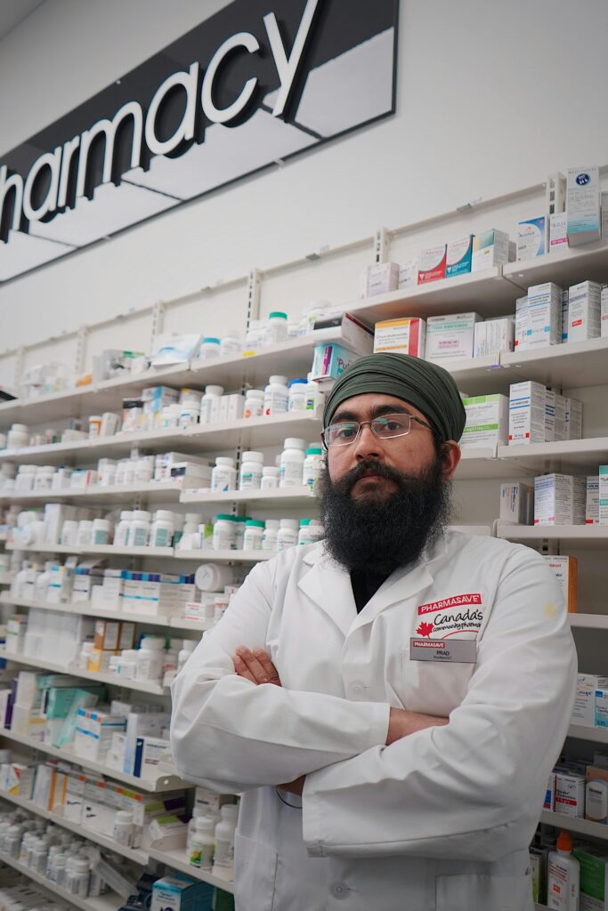 pexels-photo-14797854-14797854 Pharmacist standing with arms crossed in front of pharmacy shelves stocked with medicine.